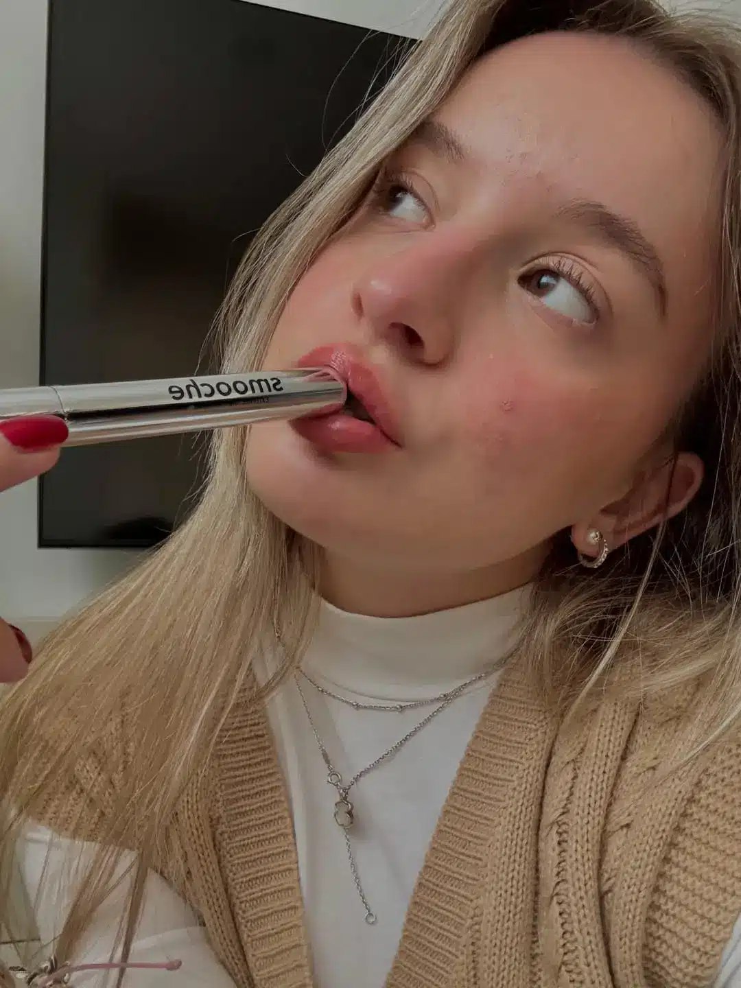 Young woman applying lipstick in cozy indoor setting with knit sweater and layered necklaces