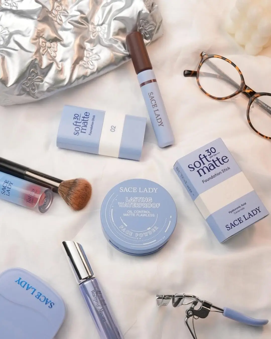 Assorted cosmetic products and tools on white fabric including face powder, foundation, and mascara